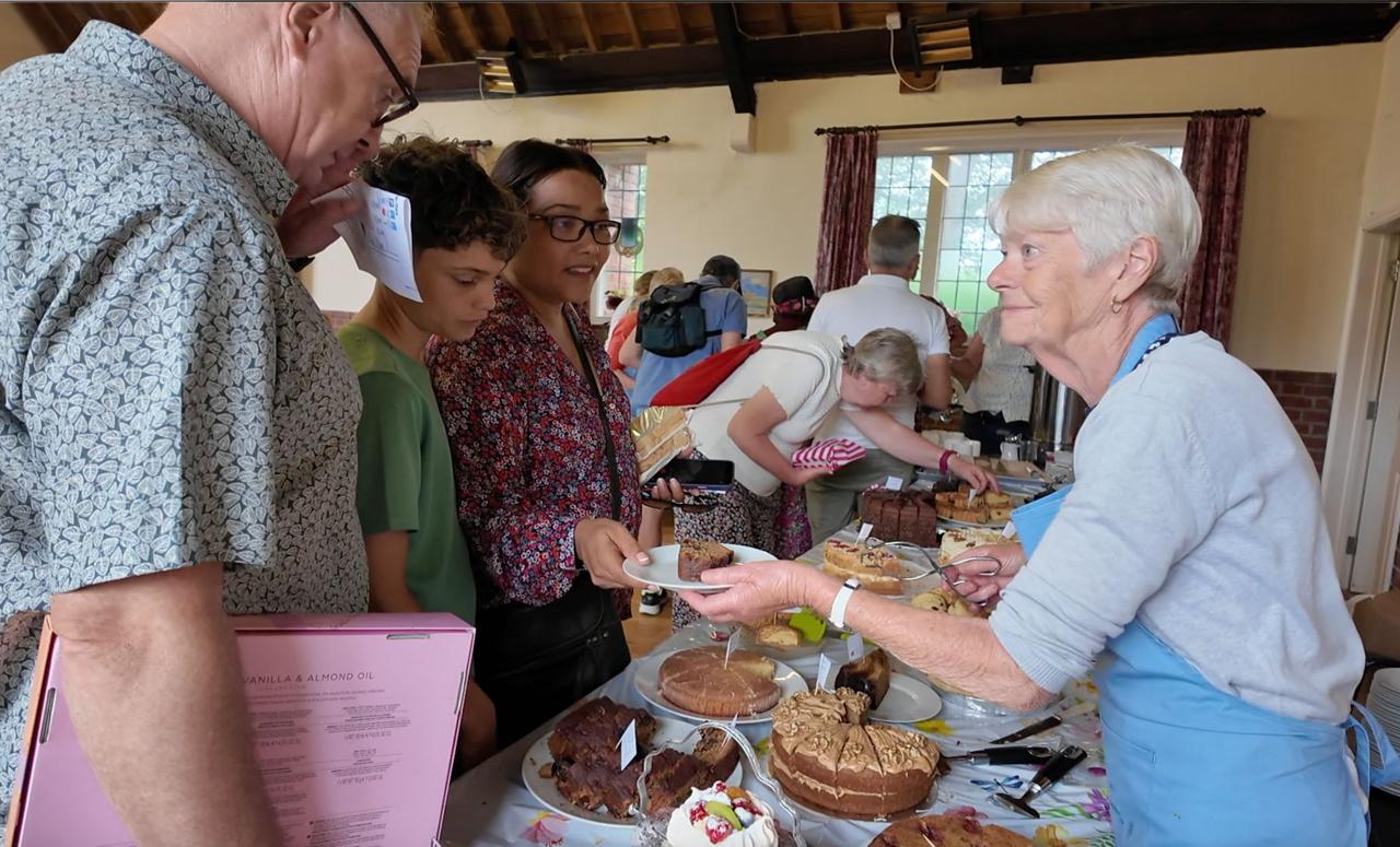 A volunteer serving visitors from a table full of homemade cakes