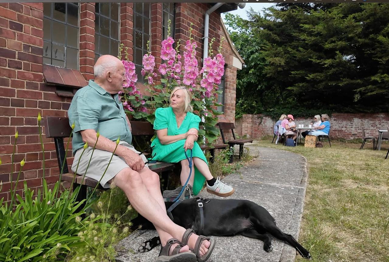 A couple with their dog on a bench beneath tall pink hollyhocks