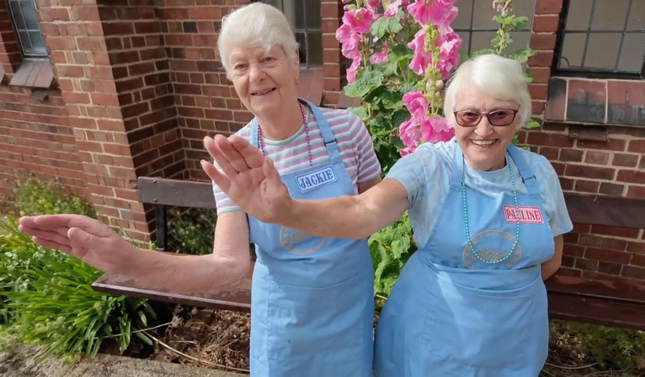 Jackie and Pauline in their aprons, waving beside the hollyhocks